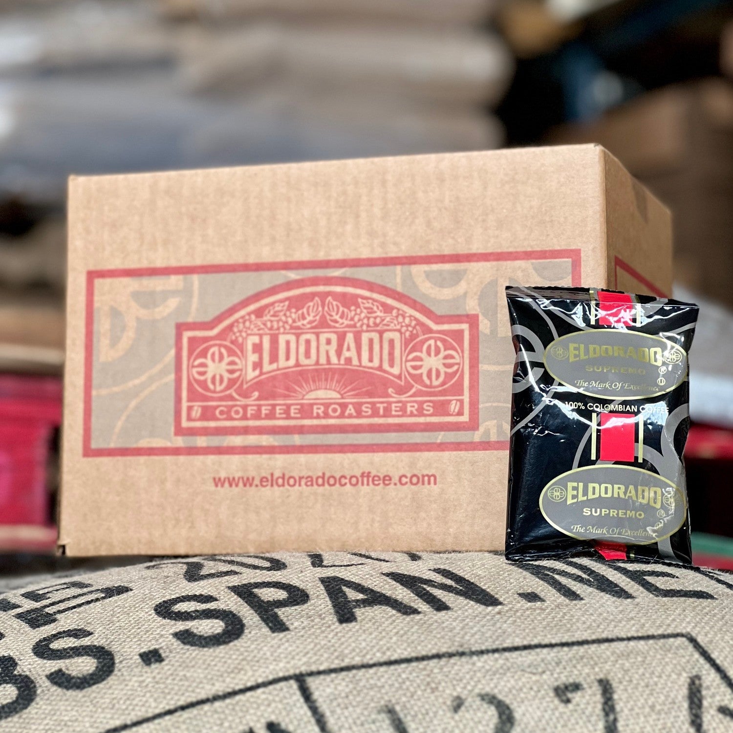 Eldorado Coffee Roasters box rests on a burlap coffee bag with a bag of Eldorado Supremo, 100% Colombian coffee, leaning against it.