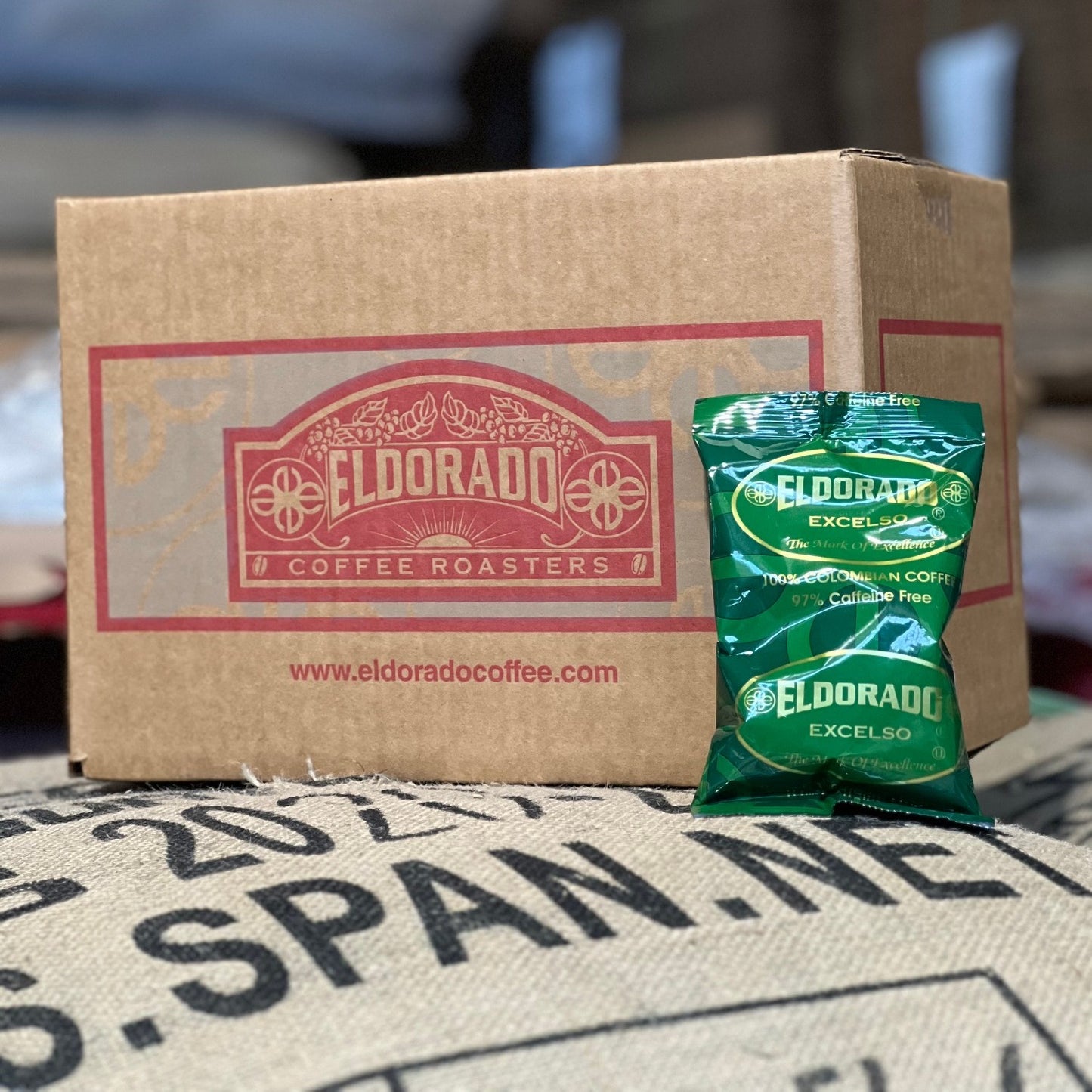 A brown cardboard box printed with the Eldorado Coffee Roasters logo sits on top of a burlap coffee bean sack. A green foil bag of Eldorado Excelso, 100% Colombian coffee in 97% caffeine free, leans against the box.